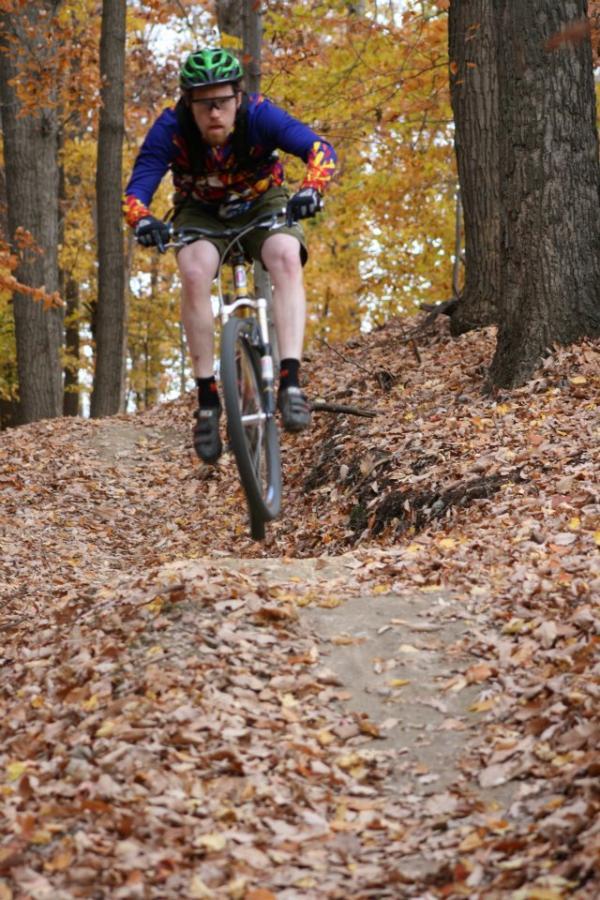 A mountain biker in a colorful jersey and helmet is airborne while jumping off a dirt ramp on a leaf-covered trail in a wooded area during fall. Laurel Hill Park mountain bike trail.