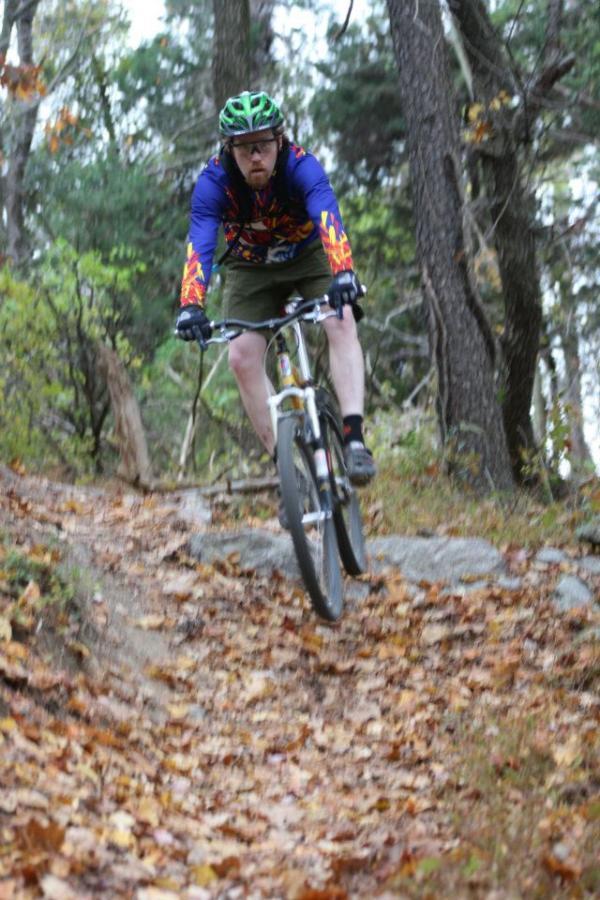 A mountain biker in colorful gear rides down a forest trail covered with autumn leaves, navigating through trees and rocks. Laurel Hill Park mountain bike trail.