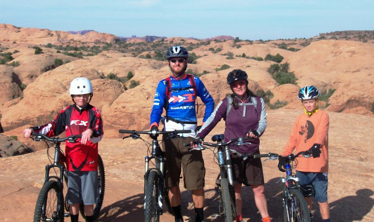 Four mountain bikers standing on rocky terrain with a desert landscape in the background. They are dressed in cycling gear and pose with their bikes. The group consists of two adults and two children. The scene captures a sunny day showcasing the adventurous spirit of biking in nature. Slickrock mountain bike trail.