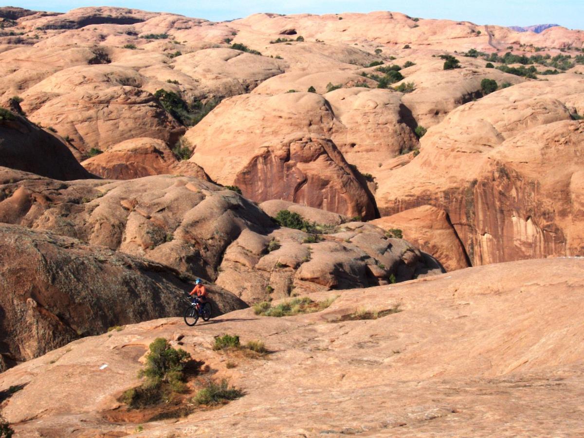 A mountain biker riding on smooth, rocky terrain surrounded by large, rounded rock formations and sparse vegetation under a clear blue sky. Slickrock mountain bike trail.