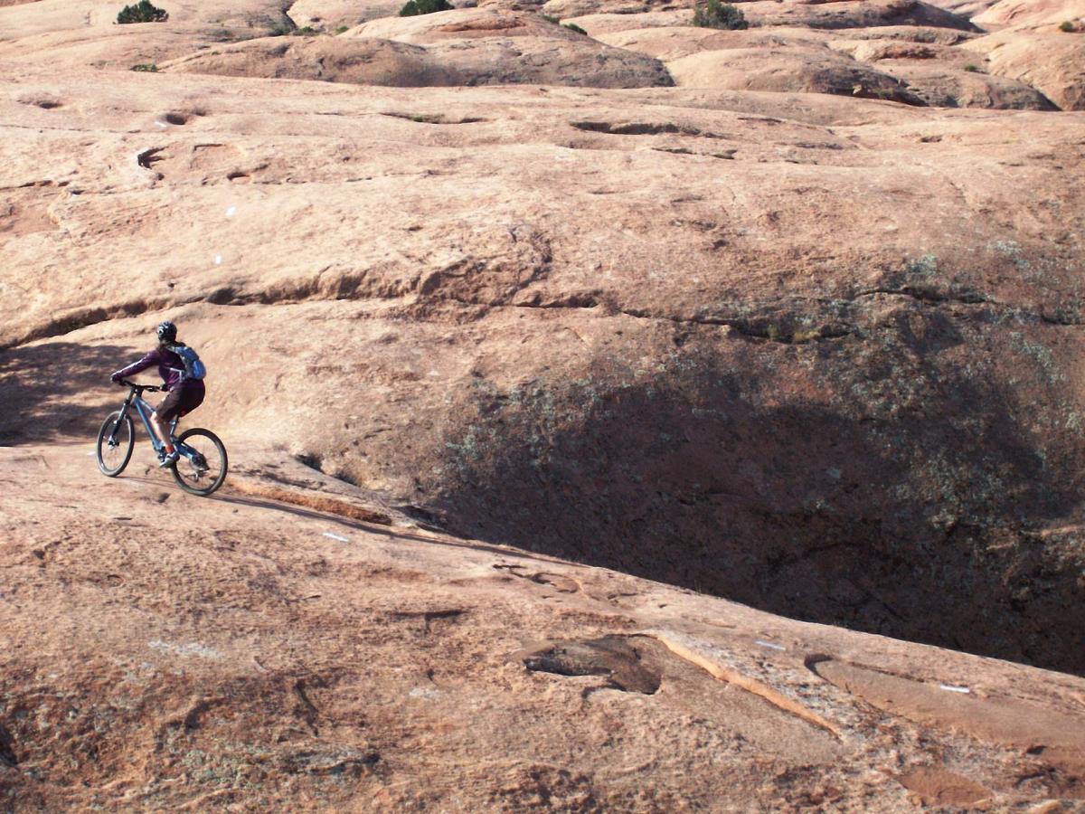 A cyclist riding on a rocky terrain, navigating over large, smooth rock surfaces with sparse vegetation in the background. The rider is wearing a helmet and a backpack, showcasing an outdoor adventure in a rugged landscape. Slickrock mountain bike trail.