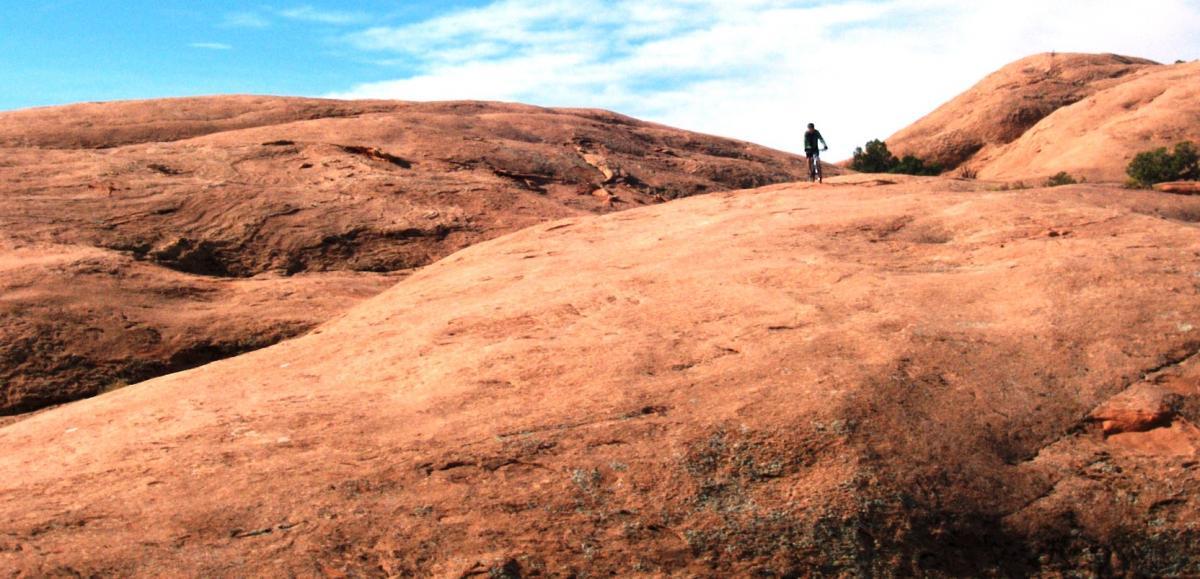 A person riding a bicycle on a rocky, desert-like terrain under a clear blue sky. The landscape features rolling hills of smooth, reddish-brown rock with patches of vegetation. Slickrock mountain bike trail.