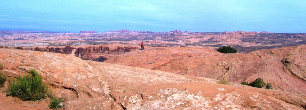 A vast desert landscape with reddish-brown rock formations and rolling hills under a clear blue sky. A lone hiker is visible on the rocky terrain, surrounded by natural scenery typical of a canyon or desert setting. Small patches of green vegetation can be seen among the rocks. Slickrock mountain bike trail.
