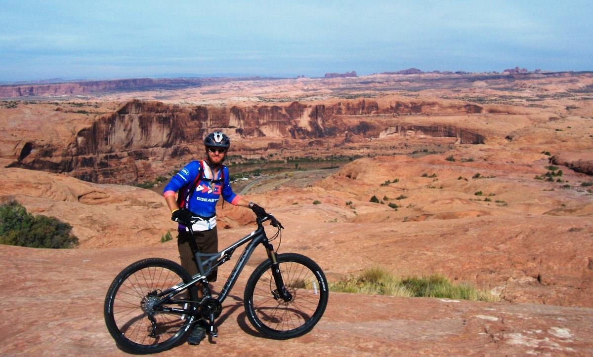 A mountain biker standing on a rocky outcrop, posing next to his bike with a vast, scenic landscape of canyons and mesas in the background. The sky is clear with some clouds, highlighting the rugged terrain and natural beauty of the area. Slickrock mountain bike trail.