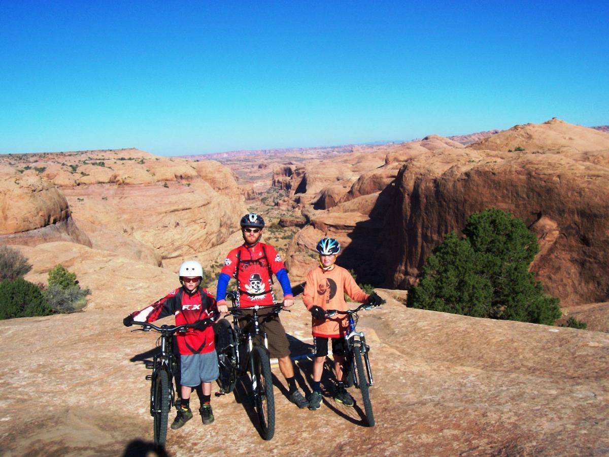 A man and two children stand on a rocky outcrop with mountain bikes, overlooking a vast desert landscape under a clear blue sky. The man is wearing a helmet and a red shirt, while the children, also in helmets, wear brightly colored shirts and shorts. The scene captures a sense of adventure and outdoor activity. Slickrock mountain bike trail.