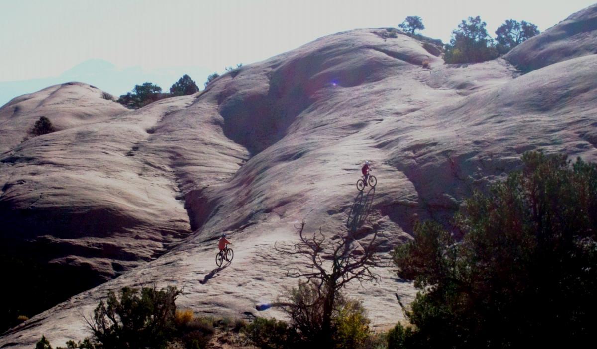 Two mountain bikers riding on a rocky terrain in a natural landscape, with trees in the foreground and a distant mountain range visible in the background. The sunlight casts shadows of the bikers on the smooth rock surface. Slickrock mountain bike trail.