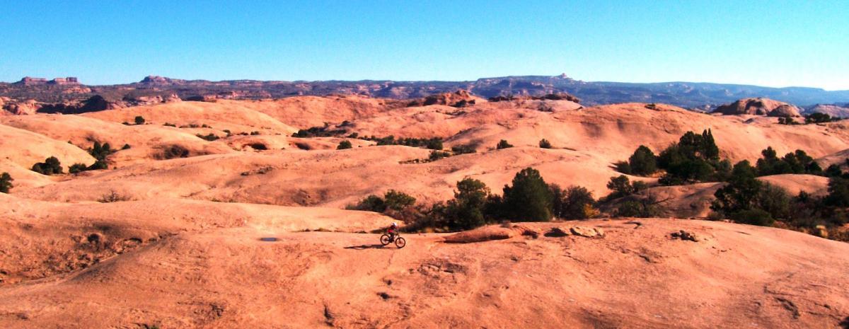 A mountain biker traverses a rocky, desert landscape under a clear blue sky, surrounded by rolling hills and sparse vegetation. Slickrock mountain bike trail.