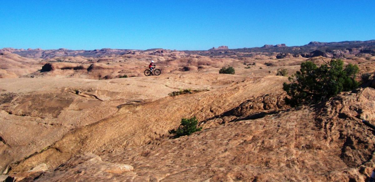 A mountain biker rides across a rocky desert landscape under a clear blue sky. The terrain features expansive, rugged rocks and sparse vegetation, typical of arid regions. Slickrock mountain bike trail.