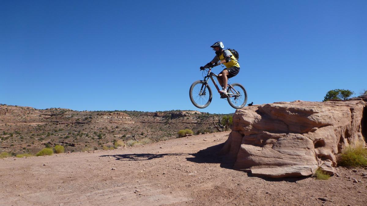 A mountain biker performs a jump over a rock formation on a dirt trail, surrounded by a dry, rocky landscape under a clear blue sky. Porcupine Rim mountain bike trail.