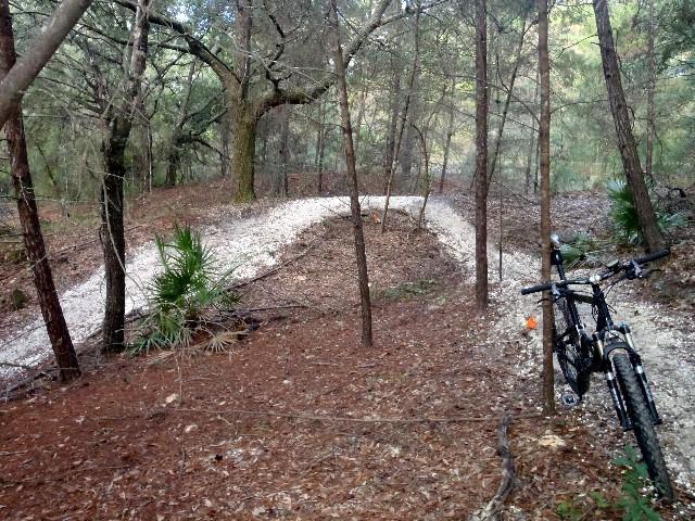 A winding dirt bike trail surrounded by trees, with a mountain bike leaning against a tree in the foreground. The trail is covered in pale gravel and leads into the dense forest, which features a mix of pine needles and foliage. Ern N Burn mountain bike trail.