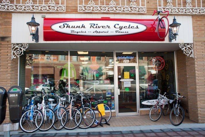 Image of a storefront for Shunk River Cycles, featuring a vibrant red sign with the store name and offerings including bicycles, apparel, and fitness equipment. Several bicycles in various colors are displayed outside the shop, showcasing a mix of styles. The entrance is welcoming, with large windows revealing a well-organized interior. Decorative elements such as a lamp post add to the storefront