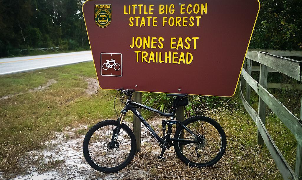 A sign for Jones East Trailhead in Little Big Econ State Forest, with a mountain bike leaning against the post. The sign features a biking symbol and is positioned near a grass area beside a road. Little Big Econ State Forest mountain bike trail.