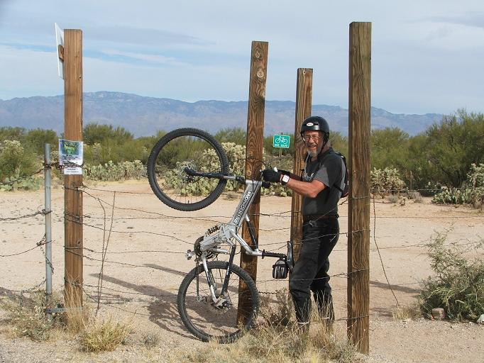 A mountain biker wearing a helmet and gloves is smiling while holding his bicycle beside a wooden fence with bike trail signs. The background features a desert landscape with mountains and vegetation. The biker has one wheel lifted off the ground. Fantasy Island mountain bike trail.