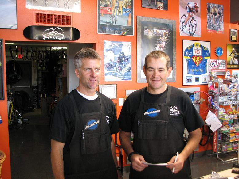 Two male bicycle shop employees stand in front of an orange wall adorned with cycling posters and memorabilia. Both are wearing black t-shirts and aprons, smiling at the camera. The shop has bicycles and cycling accessories visible in the background.