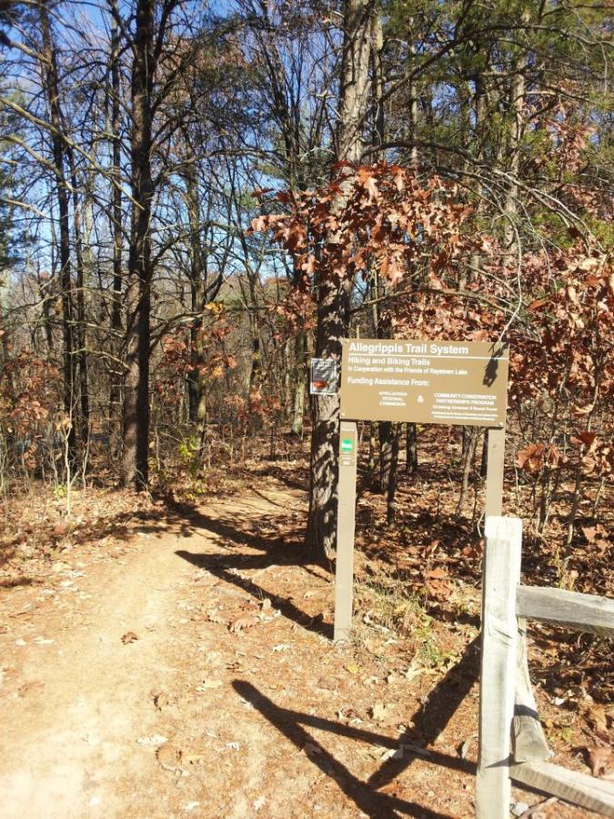 A dirt path leading into a wooded area, marked by a sign for the Allegrippis Trail System, which indicates it is for hiking and biking. The surrounding trees have autumn leaves, and a wooden fence is visible on the right side of the image. Allegrippis Trails mountain bike trail.