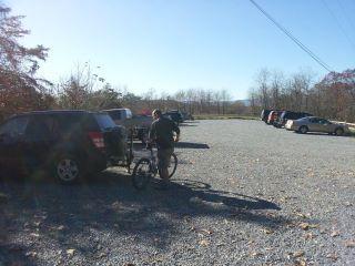 A person stands next to a bicycle in a gravel parking lot, with several parked vehicles in the background. The scene is set against a clear blue sky and trees in the distance. Allegrippis Trails mountain bike trail.