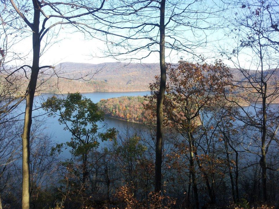 A scenic view of a lake surrounded by autumn foliage, with trees in the foreground and rolling hills in the background. The blue water reflects the colorful leaves of the trees, showcasing shades of orange, yellow, and green. The sky is clear and bright, indicating a sunny day. Allegrippis Trails mountain bike trail.