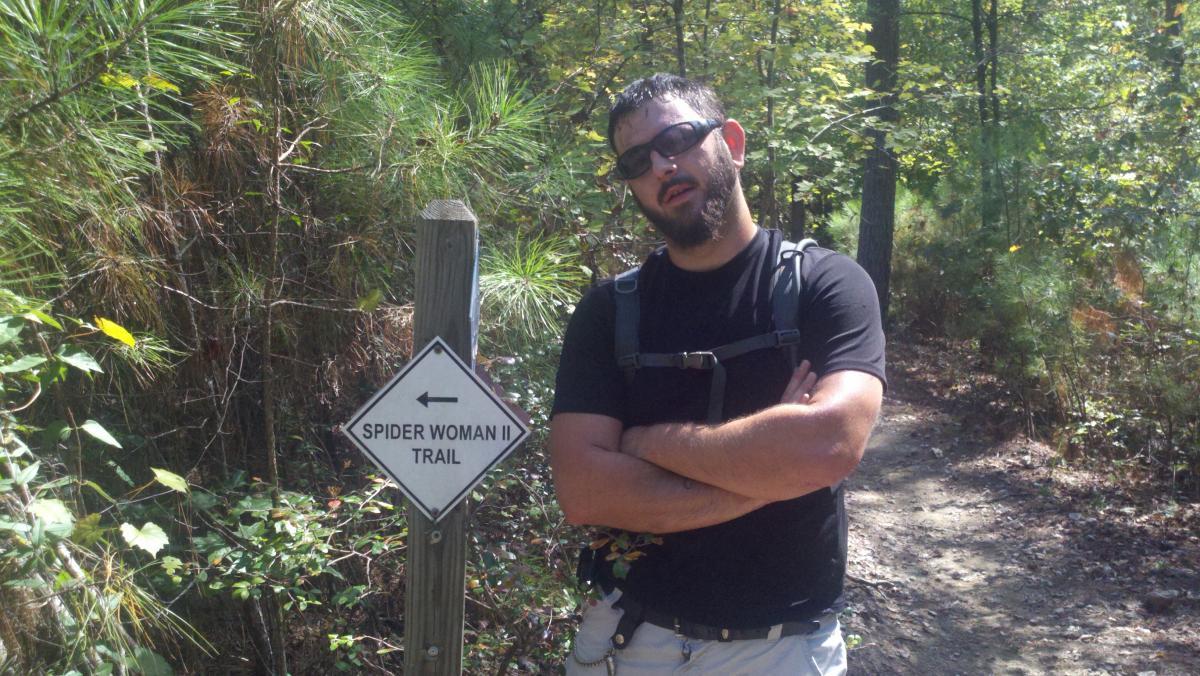 A man stands next to a trail marker indicating "Spider Woman II Trail" in a wooded area. He has a beard and is wearing sunglasses, a black t-shirt, and shorts. The background features lush greenery and trees, typical of a hiking environment. Harbison State Forest mountain bike trail.