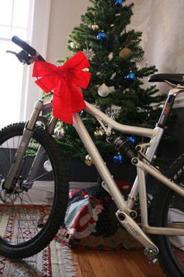 A silver mountain bike with a large red bow is positioned next to a decorated Christmas tree. The tree is adorned with blue and gold ornaments, and a colorful blanket is draped on the floor beneath it.