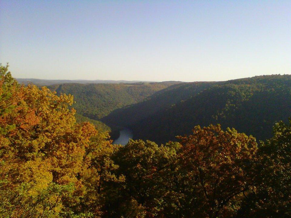 A panoramic view of a valley surrounded by rolling hills, filled with vibrant autumn foliage. A river winds through the landscape, reflecting the clear blue sky above. The scene captures the tranquility of nature during the fall season. Coopers Rock mountain bike trail.