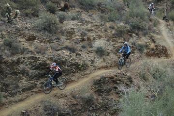 Mountain bikers navigating a winding dirt trail through a rocky landscape, surrounded by shrubs and low vegetation. Sonoran Trail mountain bike trail.