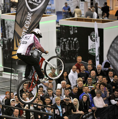 A BMX rider performs a trick in mid-air, soaring over a rail at a crowded event. The audience, comprising both men and women, watches intently, capturing the excitement. Banners and promotional materials are visible in the background, indicating a biking or sports exhibition.