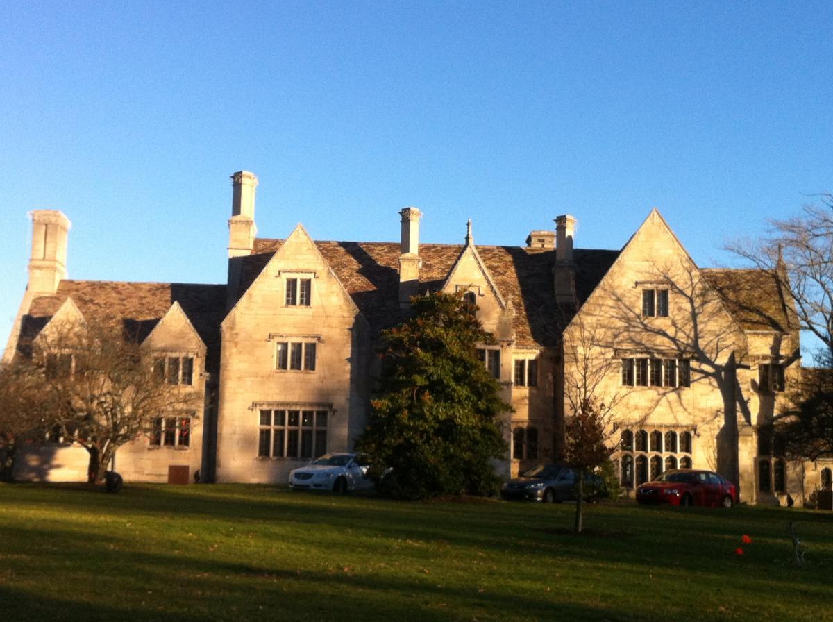 A large historic mansion with stone walls and multiple chimneys, set against a clear blue sky. The building features a variety of window styles and a sloped roof. In the foreground, there is a green lawn with several trees and parked cars. The architectural design displays elements of classic style. Hartwood Acres mountain bike trail.
