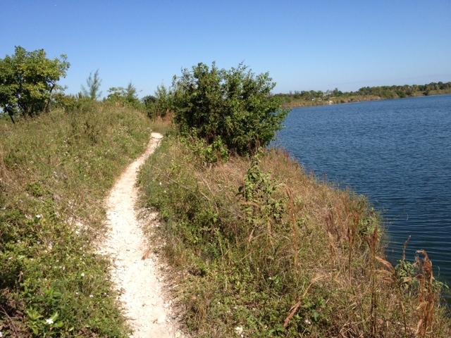 A narrow, winding dirt path bordered by lush greenery leads alongside a calm body of water under a clear blue sky. The scene captures a tranquil outdoor setting, inviting exploration and nature appreciation. Quiet Waters Park mountain bike trail.