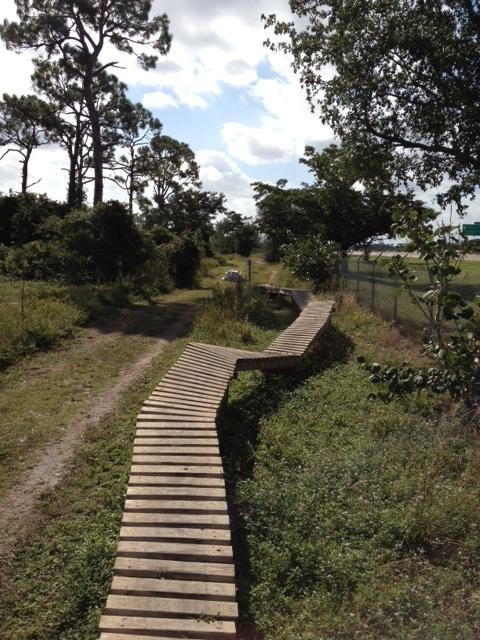 A winding wooden bridge set amidst a natural landscape, surrounded by trees and greenery, leading through a grassy trail. The sky is partly cloudy, and a fence is visible in the background. Quiet Waters Park mountain bike trail.