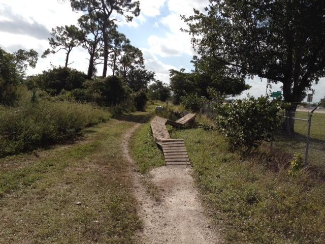 A dirt path winding through a natural landscape, flanked by shrubs and trees. A wooden plank bridge spans a section of the path, leading towards a nearby fence and signs visible in the distance. The sky is partly cloudy, suggesting a pleasant day. Quiet Waters Park mountain bike trail.