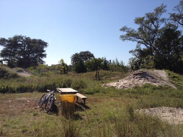 A sunny outdoor scene featuring a bicycle leaning against a wooden picnic table, with a yellow trash can nearby. In the background, there are patches of grass and trees, along with two small sandy mounds, suggesting a natural, open space. Quiet Waters Park mountain bike trail.