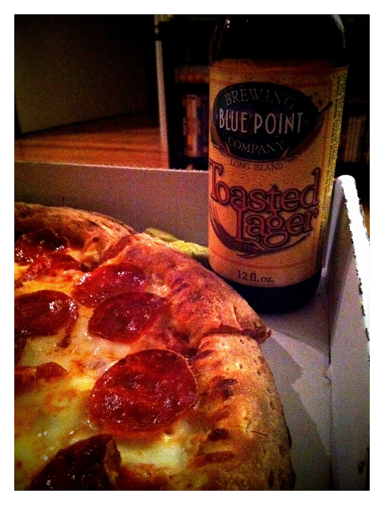 A close-up view of a pizza with pepperoni slices positioned next to a bottle of Blue Point Toasted Lager, sitting in its takeout box. The background is softly lit, suggesting a cozy indoor setting.