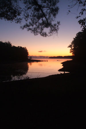 A tranquil landscape featuring a calm river at sunset, with a gradient of warm colors in the sky and silhouettes of trees along the shoreline. The water reflects the hues of the sunset, creating a serene and peaceful atmosphere.