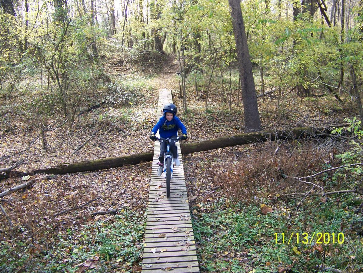 A child wearing a blue jacket and helmet rides a bicycle across a narrow wooden bridge in a wooded area filled with autumn foliage. The path is surrounded by trees and fallen leaves, creating a picturesque outdoor scene. Springhill Park mountain bike trail.