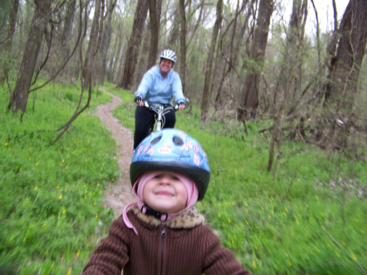 A child wearing a pink helmet and a brown sweater smiles joyfully as they ride a bike along a winding path in a green, wooded area. In the background, an adult cyclist in a blue jacket follows closely, both enjoying a day of biking outdoors. Springhill Park mountain bike trail.