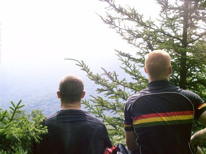 Two individuals standing side by side, viewed from behind, gazing out over a misty mountain landscape. They are surrounded by evergreen trees, and the light is soft and diffused, suggesting an early morning or late afternoon setting. The person on the left is wearing a dark shirt, while the individual on the right is dressed in a shirt with colorful stripes. Gauley Headwaters mountain bike trail.