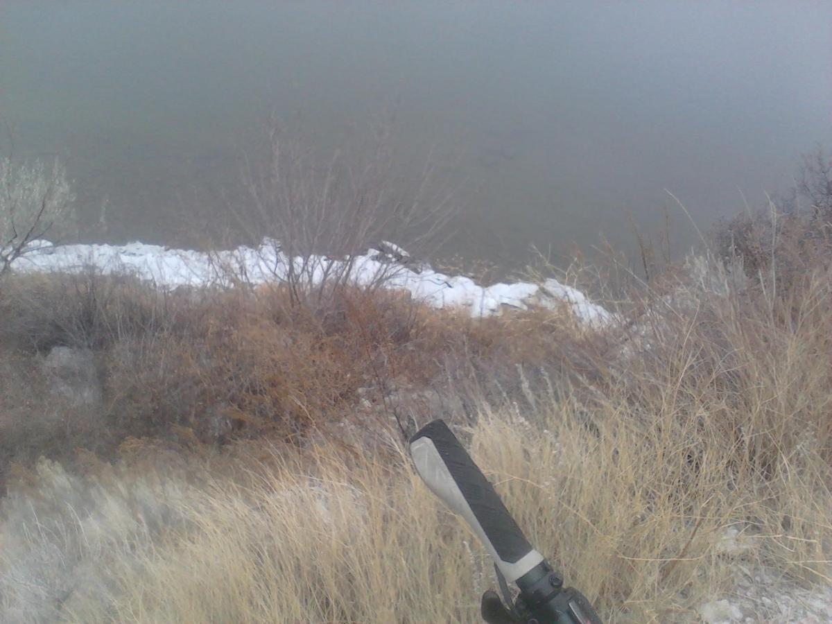 A view of a riverbank with a muddy river in the background, surrounded by dry grasses and sparse vegetation. In the foreground, a fishing rod is resting on the ground, partially obscured by tall grass and shrubs. The scene appears to be during a chilly day, with patches of white possibly indicating ice or snow along the water's edge. North Shore mountain bike trail.
