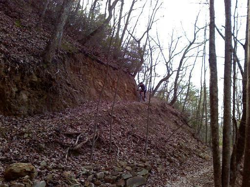 A cyclist navigating a dirt path along a hilly forest trail, surrounded by bare trees and fallen leaves. The terrain is rugged, with natural rock formations visible along the path. Five Points mountain bike trail.