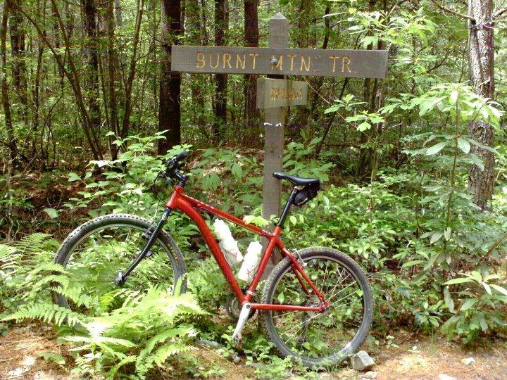 A red mountain bike leaning against a wooden trail sign marked "BURNT MTN TR" surrounded by lush green foliage and trees in a serene forest setting. Burnt Mountain Trail #11 mountain bike trail.