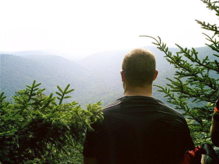 A person standing on a scenic overlook, gazing out at a mountainous landscape shrouded in mist. The foreground features green foliage framing the view, while rolling hills stretch into the distance under a bright sky. Gauley Headwaters mountain bike trail.
