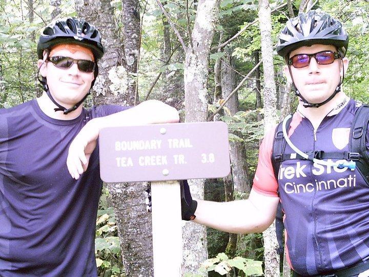 Two mountain bikers posing next to a trail sign that reads "Boundary Trail Tea Creek Tr. 3.0." They are wearing helmets and sunglasses, surrounded by trees in a forested area. One biker is dressed in a dark shirt, while the other wears a biking jersey with a logo. Gauley Headwaters mountain bike trail.