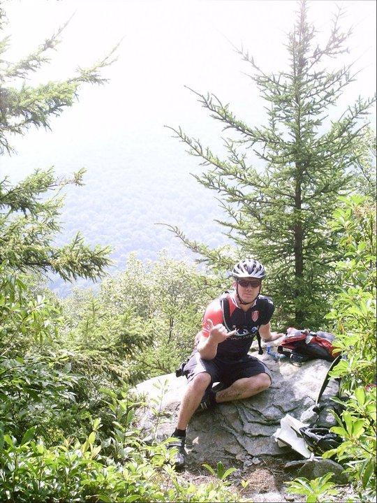 A cyclist sitting on a rock surrounded by greenery, wearing a helmet and activewear, making a hand gesture. In the background, a misty view of mountains and trees can be seen under bright daylight. Gauley Headwaters mountain bike trail.