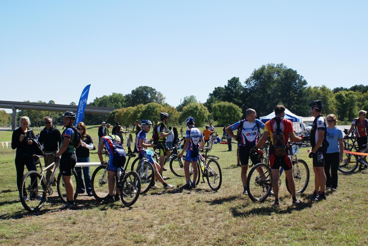 A group of cyclists wearing colorful biking jerseys stand around with their mountain bikes in a grassy area. In the background, people are gathered, and there are trees and a highway overpass. A blue flag is visible, along with tents and banners, indicating a bike event or race. The sky is clear and sunny. Springhill Park mountain bike trail.
