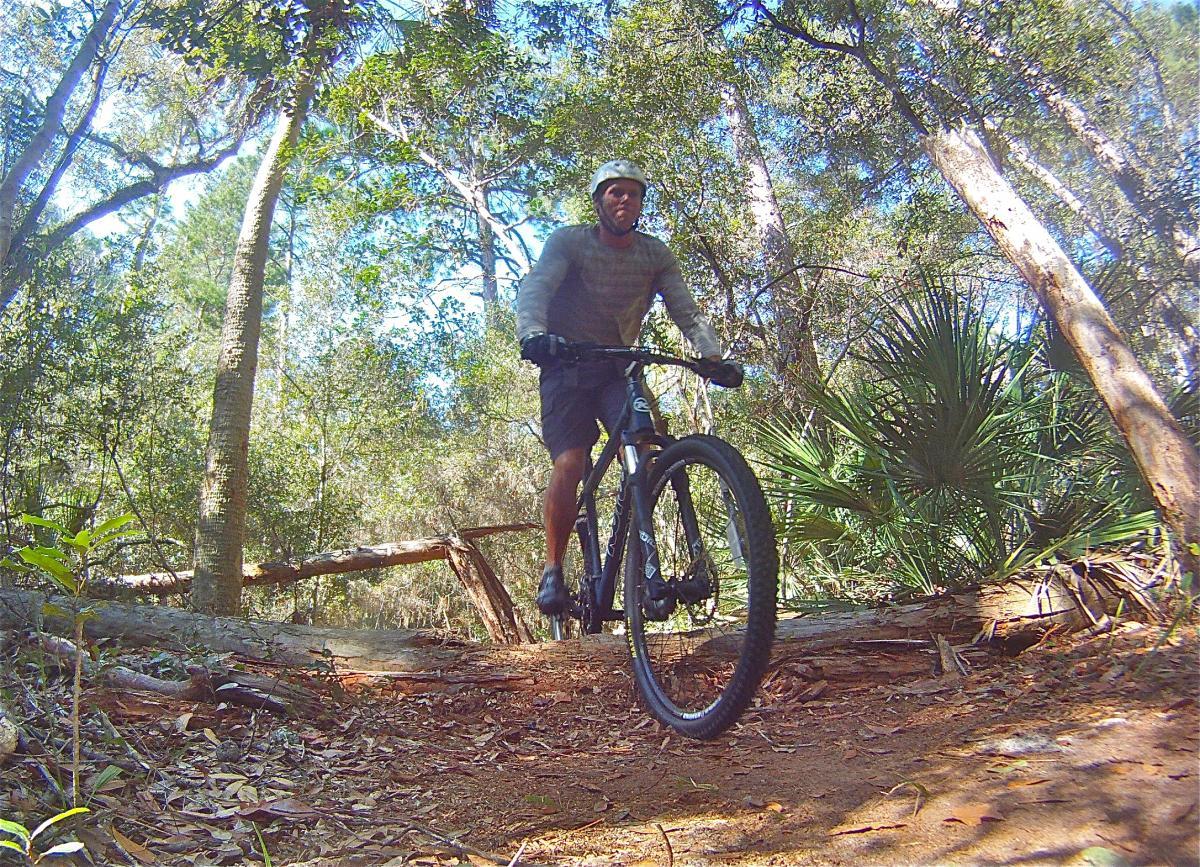 A cyclist navigating a dirt trail in a wooded area, wearing a helmet and casual outdoor attire, with trees and foliage in the background. The bike is in motion, and a fallen log can be seen on the path. Mala Compra mountain bike trail.