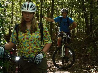 Two mountain bikers on a forest trail. One rider, wearing a colorful patterned shirt and a helmet, stands with a serious expression, while the other rider, dressed in a blue shirt and helmet, is riding a mountain bike in the background, both surrounded by dense greenery. Hobby Park mountain bike trail.