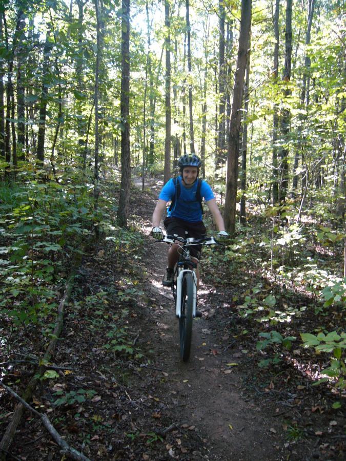 A person riding a mountain bike on a dirt trail surrounded by trees in a lush forest setting. The cyclist is wearing a blue shirt, shorts, and a helmet, and is smiling as they navigate the path. Sunlight filters through the leaves, creating a vibrant and inviting atmosphere. Hobby Park mountain bike trail.
