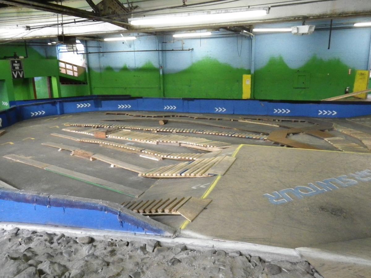 An indoor skateboarding or BMX ramp area featuring various wooden ramps and obstacles arranged on a flat surface. The walls are painted green and blue, with a sign for "First Aid" visible. The scene is well-lit with fluorescent lighting, showing a spacious environment for skateboarding or biking activities. Rays Indoor Mtb Park mountain bike trail.