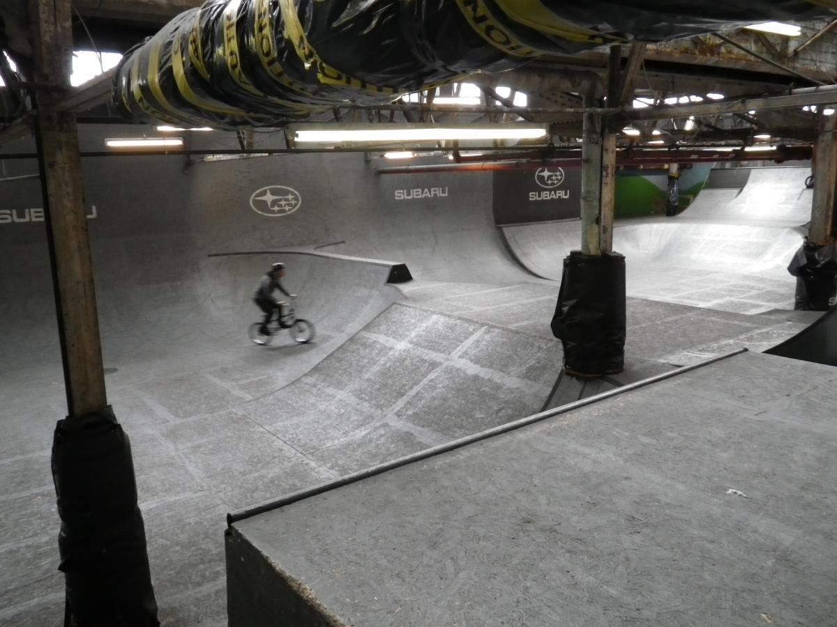 A person on a bicycle rides through an indoor skatepark featuring smooth ramps and bowl sections. The area is dimly lit with exposed wooden beams and metal structures, some areas marked with the Subaru logo. The surface is gray, and the environment has a gritty, industrial feel. Rays Indoor Mtb Park mountain bike trail.