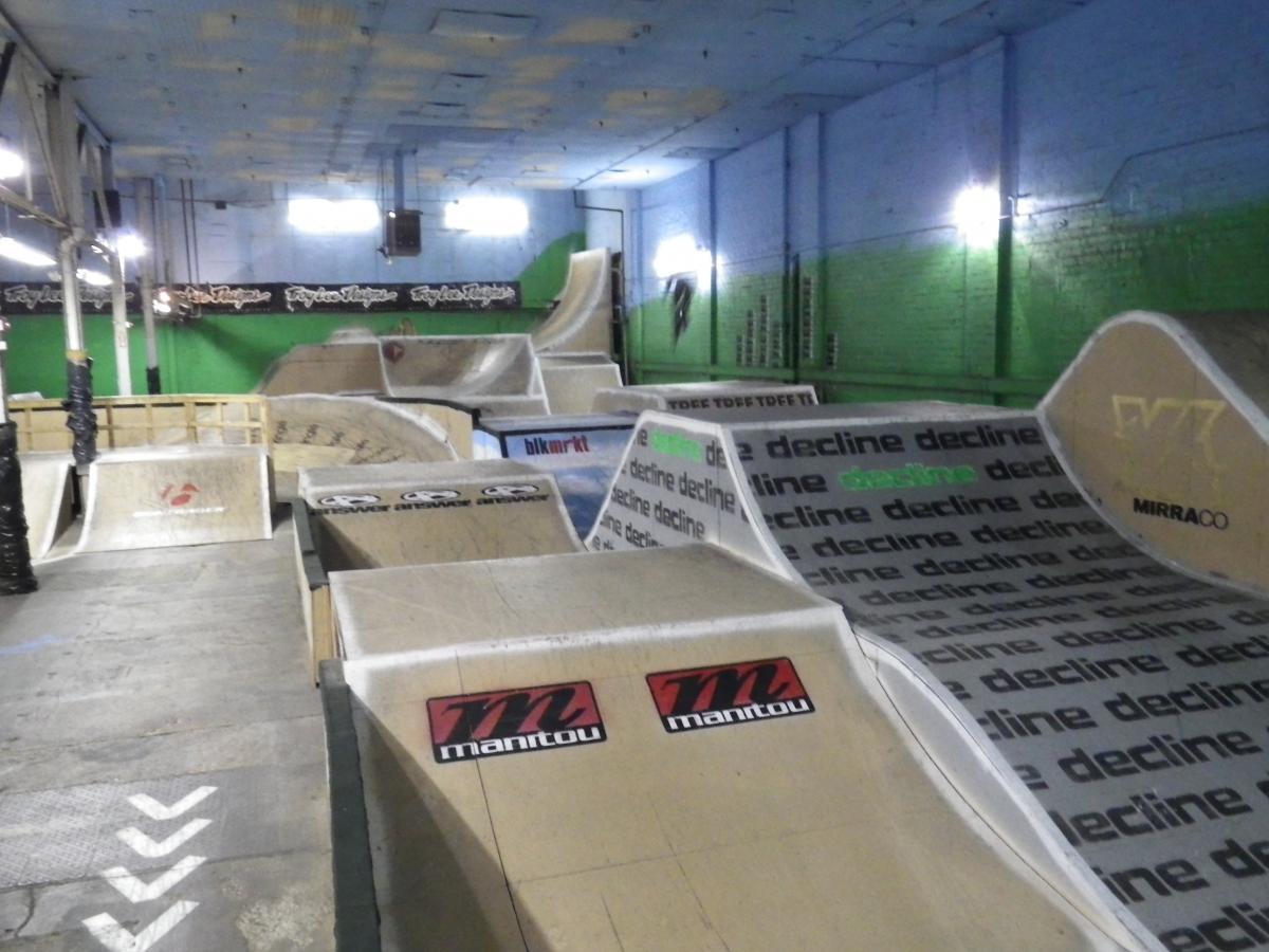 Indoor skatepark featuring various ramps and obstacles for biking and skateboarding. The walls are painted blue and green, and there are logos and branding visible on the ramps. The space is well-lit, showcasing the layout of the park, which includes a series of jumps and inclines. Rays Indoor Mtb Park mountain bike trail.