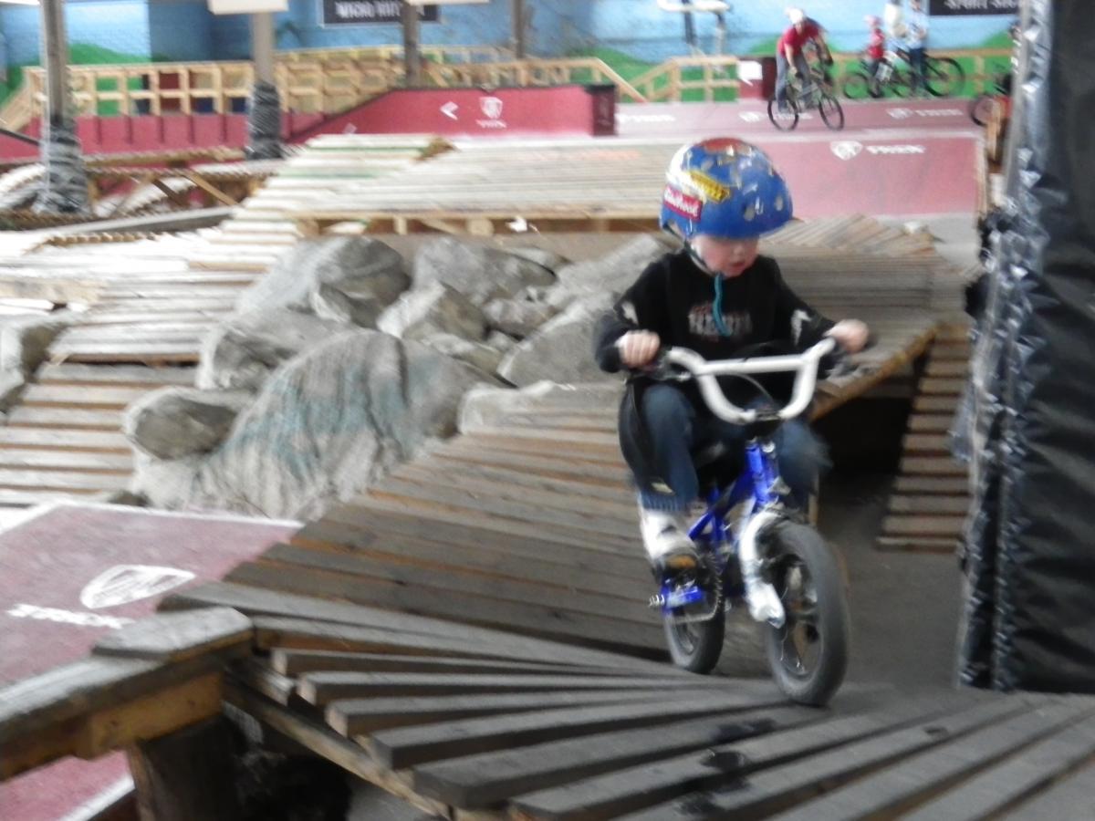 A young child riding a small blue bicycle on a wooden ramp in an indoor biking park, surrounded by rocks and other riders in the background. The child is wearing a helmet and appears focused while navigating the course. Rays Indoor Mtb Park mountain bike trail.
